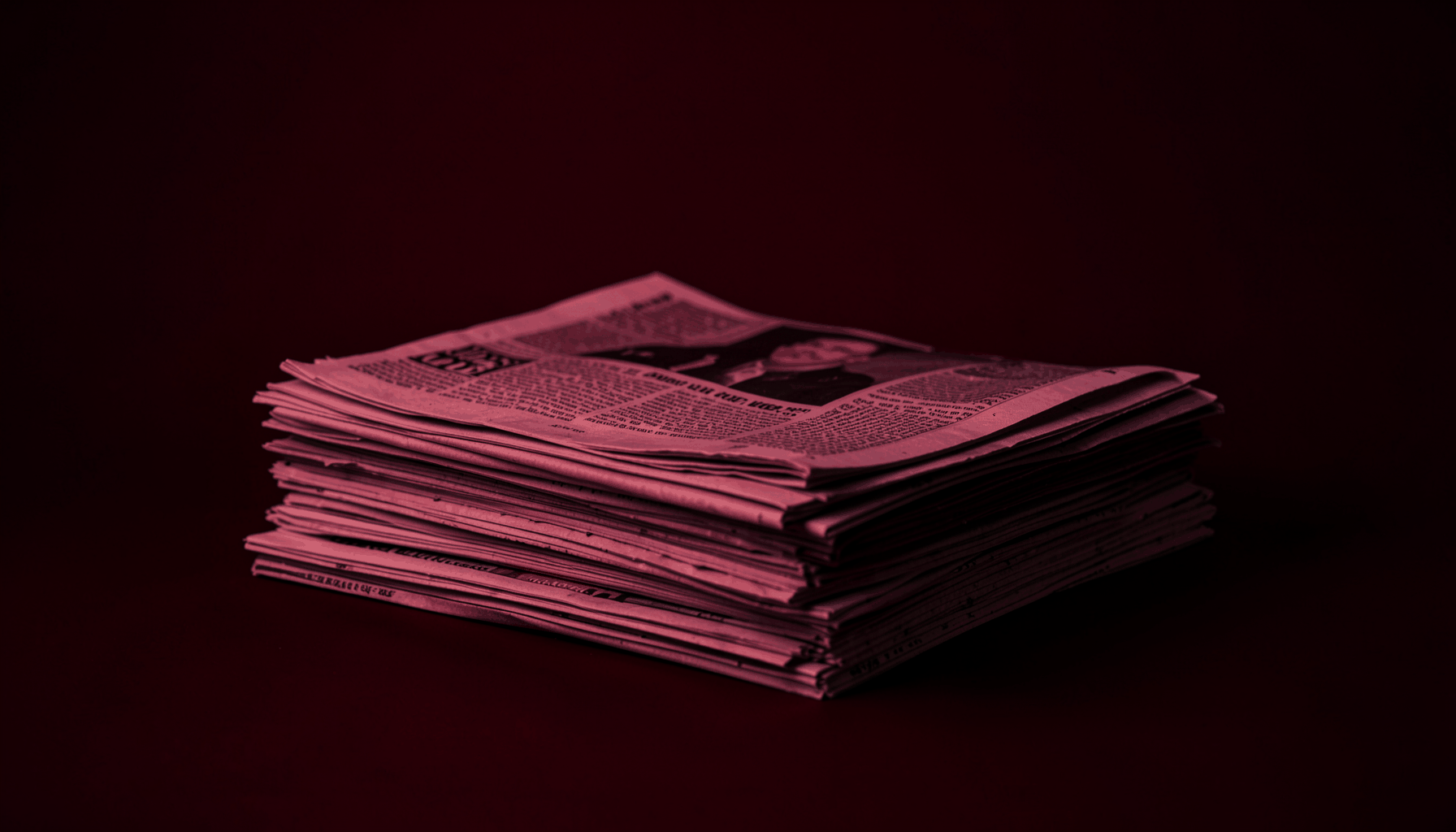 A dim newsroom-style studio photograph in deep crimson red and black tones showing a stack of folded newspapers on a dark surface